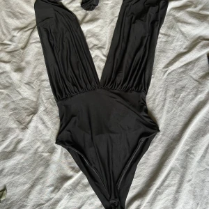 Svart baddräkt från Goddiva London - Supersnygg svart baddräkt från Goddiva London med djup v-ringning och breda axelband som kan knytas på flera olika sätt. Tillverkad i mjukt och stretchigt syntetmaterial som sitter tight och framhäver figuren. Perfekt för stranden eller poolen i sommar!
