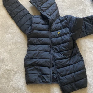 Svart pufferjacka från Lyle & Scott - Svart pufferjacka från Lyle & Scott Junior med huva och dragkedja. Jackan har det klassiska Lyle & Scott-loggan på bröstet och är tillverkad i 100% polyamid. Perfekt för kyliga dagar och har en clean, sportig look. helt ny 5 dagar gammal köpte den för 1300