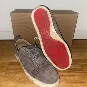 Grå mockasneakers från Christian Louboutin - Snygga grå sneakers från Christian Louboutin i mocka med klassisk röd sula. Skorna har rund tå, snörning och diskreta metallöglor på sidan för extra ventilation. Perfekt för dig som vill ha en lyxig och stilren look med en ikonisk touch.