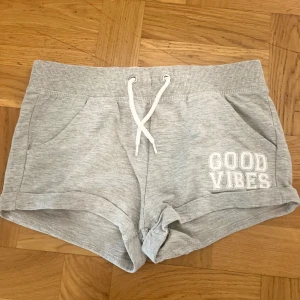 mjukisshorts - Superbekväma grå mjukisshorts med vit snörning i midjan och trycket 'GOOD VIBES' på ena benet. Shortsen har två fickor och ribbad, elastisk midja. Perfekta för chill dagar eller träning.