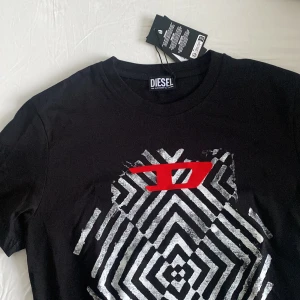 Svart t-shirt med grafiskt tryck från Diesel - Svart t-shirt från Diesel med ett fint grafiskt mönster i vitt och en röd D-logga i mitten. Klassisk rund hals och korta ärmar. Materialet är mjuk bomull som känns skön mot huden. Perfekt för dig som gillar lite mer streetstyle och tycker om att ha på dig något som få andra har. 