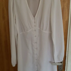 Vit långärmad klänning med knappar - Superfin vit klänning med lång ärm och djup v-ringning. Klänningen har dekorativa tygklädda knappar framtill och är gjord i ett lätt, flowy material. Perfekt för dig som gillar en stilren och elegant look. Aldrig använt SKRIV PRISFÖRSLAG!!!!