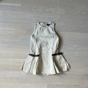 (Barn) Beige klänning från Ralph Lauren - Supersöt beige kortklänning från Ralph Lauren med ärmlös design och plisserad kjol. Klänningen har två mörka bältesdetaljer i midjan och dragkedja bak. (Barn)
