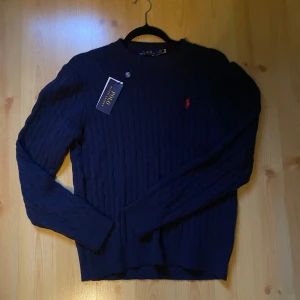 Mörkblå kabel-stickad tröja Polo Ralph Lauren - Kabelstickad mörkblå tröja från Polo Ralph Lauren med klassisk röd broderad logga på bröstet. Tröjan är i bomull, har rund hals och långa ärmar. Perfekt för dig som gillar stilren och tidlös design. Helt ny!