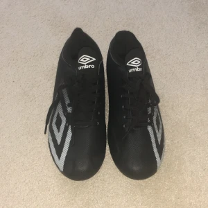 Svarta Umbro fotbollsskor med snörning - Snygga svarta fotbollsskor från Umbro med klassisk vit logga på sidan och tungan. Skorna har snörning och är tillverkade i syntetmaterial för en lätt känsla på planen. Perfekta för dig som vill ha stilrena och funktionella skor till match eller träning.