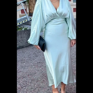 Ljusgrön långklänning med v-ringning - Superfin ljusgrön långklänning från gina tricot i glansigt satinliknande material. Klänningen har djup v-ringning, ballongärmar med resår vid handlederna och markerad midja. Perfekt för dig som vill sticka ut med en elegant och trendig look.