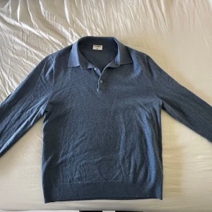 Blå pikétröja i ull från Filippa K - Snygg blå långärmad pikétröja från Filippa K med klassisk krage och knäppning framtill. Tröjan är tillverkad i mjuk ull och har en stilren, minimalistisk look som passar perfekt till jeans eller chinos.