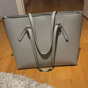 Grå axelväska med dragkedja - Stilren grå axelväska i syntetmaterial med guldiga metalldetaljer och lång rem. Väskan har en fyrkantig form, dragkedja upptill och ett rymligt innerfack med extra ficka. Perfekt för att bära med sig laptop, böcker eller andra nödvändigheter.