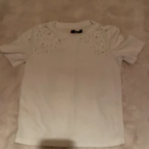 Vit T-shirt  med pärlor och stenar - Superfin vit T-shirt i strukturerat tyg med korta ärmar. Dekorerad med pärlor och glittrande stenar över axlar och bröst, vilket ger en lyxig och trendig känsla. Perfekt för dig som vill sticka ut lite extra! Rund halsringning och rak passform.