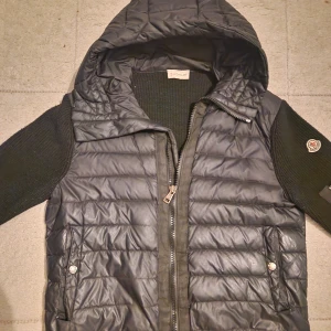 Moncler cardigan - Storlek M och S borde passa Snygg svart dunväst från Moncler med quiltad framsida, stickade svarta ärmar och huva. Västen har dragkedja framtill och två fickor med tryckknappar. Perfekt för lager-på-lager och streetstyle. Pris kan diskuteras 