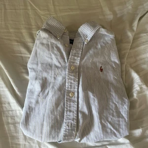 Randig skjorta från Ralph Lauren - Snygg skjorta från Ralph Lauren i vitt och blått med klassiskt randigt mönster. Skjortan har knappar framtill, button-down krage och den ikoniska broderade polospelaren på bröstet. Tillverkad i 100% bomull för en skön och luftig känsla. Perfekt att matcha till linnebyxorna i min profil.