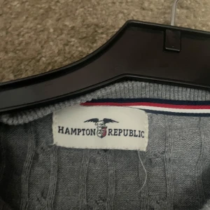 Grå kabelstickad tröja Hampton Republic - Kabelstickad grå tröja från Hampton Republic med broderad logga på bröstet. Klassisk modell med rund hals och långa ärmar, perfekt för lager-på-lager. Mjuk och skön känsla, passar till jeans eller chinos.