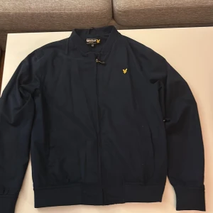 Mörkblå jacka från Lyle & Scott - Snygg mörkblå jacka från Lyle & Scott med klassisk gul logga på bröstet. Jackan har dragkedja framtill, ribbade muddar och krage. Perfekt för dig som vill ha en stilren och enkel jacka med sportig känsla. Nyskick,använd ett fåtal gånger,