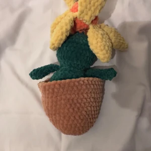 Stickad figur  - Bilderna visar en virkad eller stickad mjukisleksak som föreställer en blomma i en kruka, inte en bok. Ingen information om boktitel, författare eller innehåll kan identifieras från bilderna.