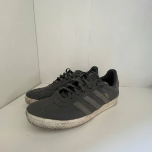 Adidas Gazelle  - Klassiska Adidas Gazelle sneakers i mörkgrått skinn med de ikoniska tre ränderna på sidan. Låg modell med snörning och platt sula. Guldtexten 'GAZELLE' på sidan ger en snygg detalj. Perfekta för en clean och sportig stil.