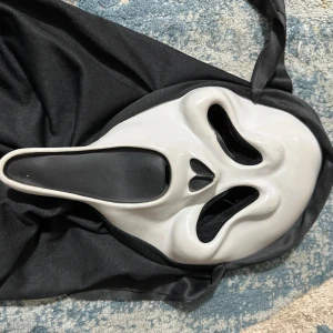 Vit och svart Scream-mask - Säljer en ikonisk Scream-mask i vit och svart plast med tillhörande svart tyg som täcker huvudet. Perfekt för att sticka ut på maskerad eller halloween. Masken har de klassiska utdragna ansiktsdragen och täcker hela ansiktet.