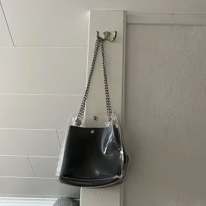 Svart axelväska med kedjerem - Snygg svart axelväska från asos med en genomskinlig  ytterväska och kedjerem i metall. Den svarta väskan inuti går att ta ut. Säljer då jag tyvärr knappt använder den.