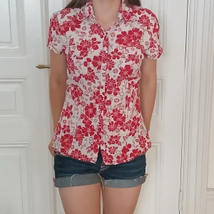 Rödblommig kortärmad skjorta - Snygg kortärmad skjorta med rött blommönster på vit botten. Skjortan har klassisk krage, knappar framtill och två bröstfickor. Perfekt för sommarens varma dagar och ger en fräsch vibe till din outfit. Står L men skulle säga att den passar någon mellan xs-m beronde på hur tajt man vill att den ska sitta, jag på bilden bär ofta xs/s. Köpt på en second hand men har endast andvänt ett fåtal gånger så den är i bra skick❤️ 