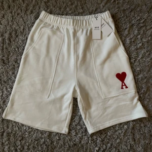 Vita shorts från Ami Paris med hjärtdetalj - Snygga vita shorts från Ami Paris med elastisk midja och två stora fickor framtill. På vänster ben finns en röd broderad hjärtdetalj med bokstaven A. Tillverkade i mjuk bomull, perfekta för sommardagar. Enkel och clean design som sticker ut med den ikoniska loggan.