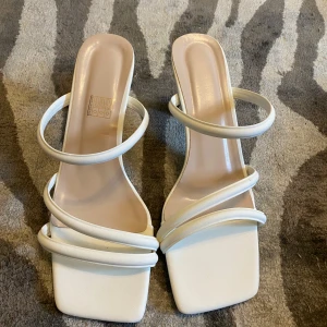Vita sandaler med klack och remmar - Trendiga vita sandaler med fyrkantig tå, flera tunna remmar och unik geometrisk klack. Snygg minimalistisk design som passar perfekt till sommarens outfits. Tillverkade i syntetmaterial med platt sula framtill och låg klack.