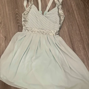 Ljusblå kortklänning med paljettdetaljer - Superfin ljusblå kortklänning med omlottliv och smala axelband. Klänningen har glittriga silverpaljetter längs midjan och sidorna, samt ett plisserat liv. Kjolen är lätt och luftig med vacker fall. Perfekt för dig som vill sticka ut med lite extra bling! Går strax över knän för mig som e 164