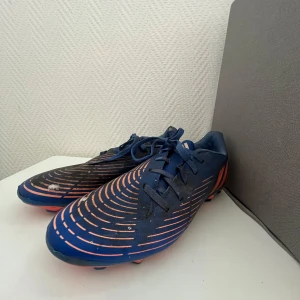 Adidas fotbollskor - Snygga fotbollsskor från Adidas i blått med orangea detaljer och mönster. Skorna har snörning och plös med låg profil, samt orange dobbar för bra grepp på planen. Tillverkade i syntetmaterial och designade för att sticka ut på matchen.
