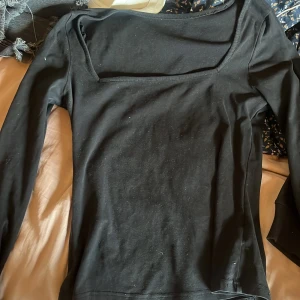 Svart långärmad topp med fyrkantig ringning - Svart långärmad topp med snygg fyrkantig ringning som ger en modern vibe. Tight passform som framhäver figuren och passar perfekt till jeans eller kjol. Tillverkad i mjukt bomullsmaterial som känns skönt mot huden.