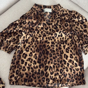 Leopardmönstrad blus från Neo Noir - Snygg blus från Neo Noir i leopardmönster med bruna och svarta toner. Blusen har korta puffärmar med resår vid ärmslut och en v-ringad halsringning. Materialet är glansigt och känns lyxigt, perfekt för dig som vill sticka ut.
