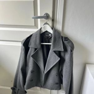 Grå croppad skepparkavaj från H&M - Snygg grå skepparkavaj från H&M med dubbelknäppning, breda slag och axelklaffar. Jackan är croppad och har stora knappar samt dekorativa detaljer vid ärmsluten. Perfekt för dig som vill ha en stilren och trendig look.