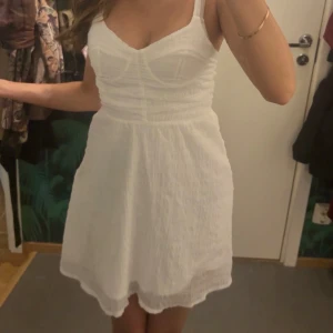 Vit kort sommarklänning med smock - Superfin vit kort klänning med smockad överdel och markerad byst. Axelbandsmodell med lätt volang i kjolen, perfekt för varma dagar. Tunn och luftig, ger en fräsch och söt look.