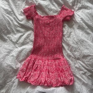 Rosa kortklänning med mönster - Supersöt rosa kortklänning med vita mönster och smockad överdel. Klänningen har korta ärmar och en volangig kjol. Använd 1 gång så är som ny🩷🩷