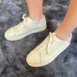 Vita sneakers från Axel Arigato - Stilrena vita sneakers från Axel Arigato med diskret logga på sidan och häl i ljusgrå mocka. Skorna har snörning, platt sula och är tillverkade i skinn med detaljer i mocka. Perfekta för en clean och modern look. Nypris 2500kr