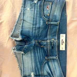 Blå jeansshorts från Hollister - Snygga blå jeansshorts från Hollister med råa, fransiga kanter och slitna detaljer. Klassisk femficksmodell med orangea sömmar och logga på bakfickan. Perfekta för varma dagar när du vill ha en avslappnad och trendig look.