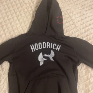Svart HOODRICH hoodie med tryck - Svart hoodie från HOODRICH med stor vit logga och duvor på ryggen. På framsidan finns trycket 'From Nothing To Something' i rött och vitt. Klassisk huva och känguruficka, perfekt för en streetwear-look.