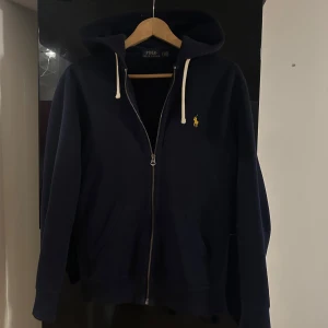 Mörkblå hoodie från Polo Ralph Lauren - Mörkblå hoodie från Polo Ralph Lauren med dragkedja, huva med vita snören och klassisk gul broderad logga på bröstet. Tröjan är i storlek S och är i bra skick. 