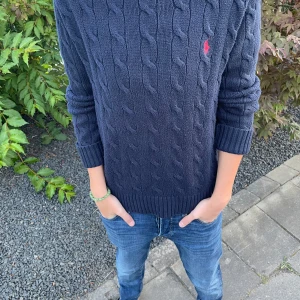 Mörkblå stickad tröja Polo Ralph Lauren - Säljer en mörkblå kabelstickad tröja från Polo Ralph Lauren med klassisk röd logga broderad på bröstet. Tröjan har rund hals, ribbade muddar och är långärmad. Perfekt för dig som gillar stilren och tidlös design. Storlek s