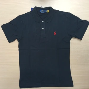 Svart pikétröja från Polo Ralph Lauren M - Svart pikétröja från Polo Ralph Lauren med klassisk krage och två vita knappar. Röd broderad logga på bröstet ger en snygg kontrast. Tröjan är kortärmad och gjord i mjuk bomull som känns skön mot huden. Perfekt för en clean och stilren look. Sprillans ny