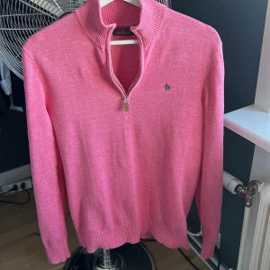 Rosa half zip tröja från Morris - Säljer en snygg rosa tröja från Morris med half zip och ribbad krage. Tröjan har ett litet broderat emblem på bröstet och är tillverkad i mjukt stickat material. Perfekt för dig som gillar färg och vill ha en stilren look.