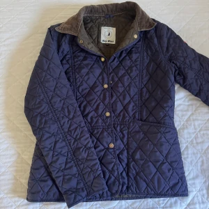Quiltad jacka från Key West, marinblå - Quiltad marinblå jacka från Key West med bruna detaljer på kragen. Jackan har tryckknappar framtill, två stora fickor och ett klassiskt rutmönster. Perfekt för dig som gillar stilren och tidlös look. Fodrad insida och långärmad modell.