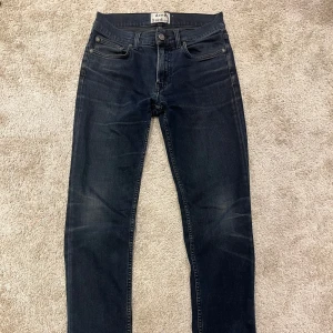 Acne Studios Ace jeans blå/svart 29/32 (slim fit) - Snygga Acne Studios Ace jeans i en mörkblå till svart tvätt. Klassisk femficksmodell med raka ben och normal passform. Materialet är 94% bomull, 5% polyester och 1% elastan för lite stretch. Perfekta för dig som gillar stilrena och tidlösa jeans.