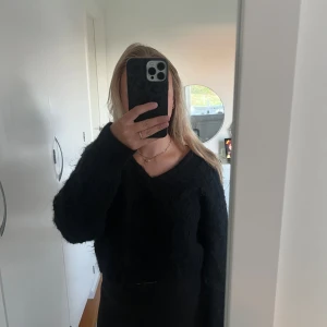 Svart fluffig stickad tröja - Mysig svart stickad tröja med fluffig känsla och v-ringad hals. Tröjan har lång ärm och en lite croppad passform som ger en trendig look. Perfekt att styla med jeans eller kjol för en chill vibe.