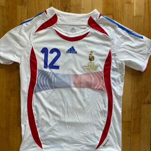 Frankrike Henry 12  - Säljer en officiell Frankrike fotbollströja från Adidas med Henry och nummer 12 på ryggen. Tröjan är vit med röda och blå detaljer, samt broderad fransk landslagslogga och VM 2006-märke. Korta ärmar och tillverkad i lätt funktionsmaterial.