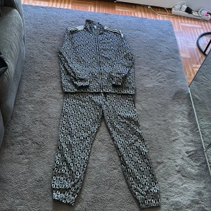 Svartvit mönstrad träningsväst - Säljer en svartvit träningsväst från Fendi  med heltryckt Fendi-logga i rutmönster. Västen har dragkedja framtill, ståkrage och klassiska svart vita ränder på axlarna. Perfekt för dig som gillar statement-plagg och sportig stil.