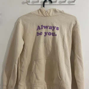 Beige hoodie med lila text från H&M - Beige hoodie från H&M med lila texten 'Always be you.' på bröstet. Tröjan har huva, magficka och är långärmad. Tillverkad i mjuk bomullsmix som är perfekt för kyligare dagar. Enkel och stilren design med en peppig touch.