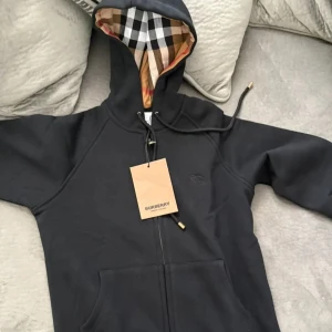 Svart Burberry hoodie med dragkedja - Svart hoodie från Burberry med klassiskt rutigt foder i huvan och gulddetaljer på snörningen. Tröjan har dragkedja framtill och fickor. Perfekt för dig som vill ha en stilren och lyxig look med ikoniska Burberry-detaljer. HELT NY!!! 