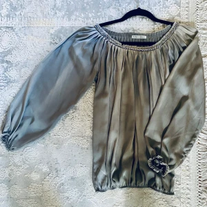 Grå blus med pärldetaljer från Ambre Babzoe - Supersnygg grå blus från Ambre Babzoe i glansig polyester. Vida långa ärmar med resår vid handlederna och plisserad design. Halsringningen är dekorerad med pärlor och små stenar för extra bling. Perfekt för dig som gillar statement-plagg! Nypris ca 1600 kr. 