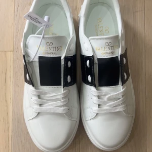 Valentino Garavani vita sneakers - Snygga vita sneakers från Valentino Garavani med bred svart läderdetalj över snörningen och ikoniska nitar baktill. Skorna har rund tå, platt sula och klassisk snörning. Perfekta för dig som gillar stilrena och lyxiga detaljer.