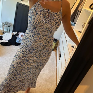 Blommig långklänning med knyt - Supersöt långklänning med blått blommönster på vit botten. Smala axelband och knytning vid bysten.