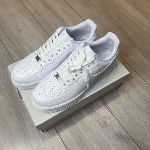 Nike Air Force 1 helvita sneakers - Klassiska Nike Air Force 1 sneakers i helvitt skinn med perforerad tå, rund tå och platt sula. Snygg och tidlös modell med snörning och diskret Nike-logga på hälen. Perfekta för dig som vill ha en clean och stilren look.                                     Storlek:43  Skick: Helt nya  📦 Skickas inom 1–3 dagar. 📩 För mer information eller frågor, kontakta mig gärna.