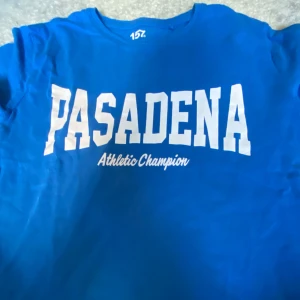 Blå Pasadena t-shirt från lager 157 - Blå t-shirt från lager 157 med stort vitt tryck 'PASADENA Athletic Champion' på bröstet. Klassisk rund halsringning och korta ärmar. Tillverkad i 100% bomull för en skön och luftig känsla. Perfekt för dig som gillar sportig och avslappnad stil.